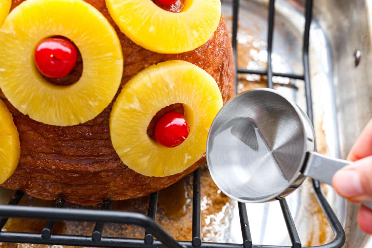 Brown Sugar Pineapple Glazed Ham is coated with a sweet and tangy spiced glaze that caramelizes on the edge of the ham. It's covered in juicy pineapple rings and maraschino cherries that add a tangy sweetness and juices to the succulent ham. Close up shot of someone pouring water into the bottom of the roasting pan.