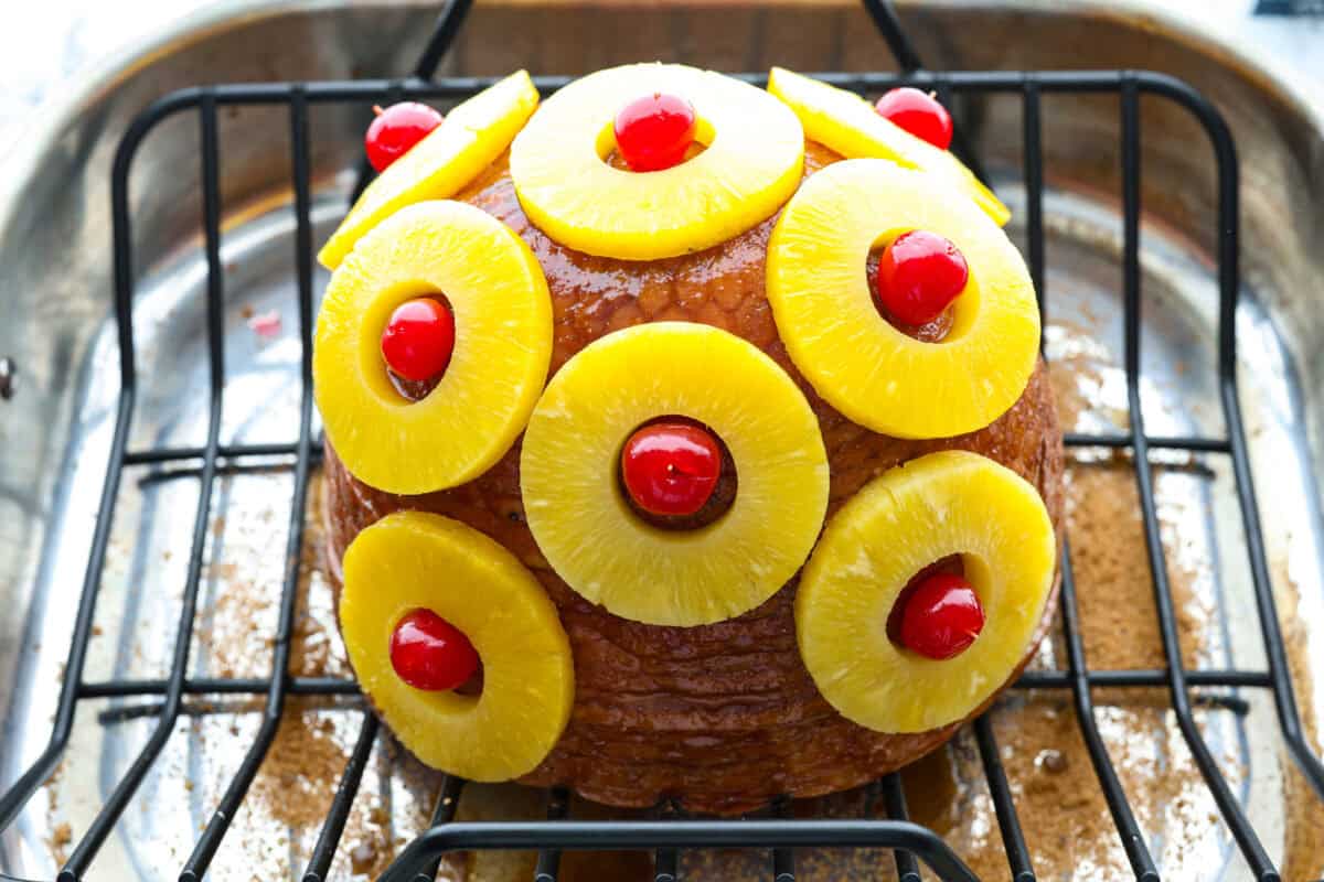 Brown Sugar Pineapple Glazed Ham is coated with a sweet and tangy spiced glaze that caramelizes on the edge of the ham. It's covered in juicy pineapple rings and maraschino cherries that add a tangy sweetness and juices to the succulent ham. Angle shot of the ham covered in pineapple rings and cherries.