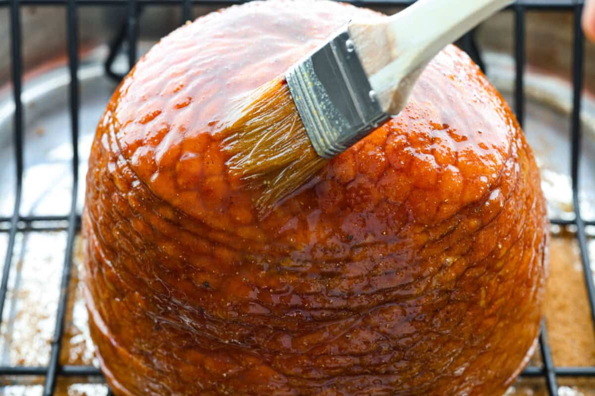 Brown Sugar Pineapple Glazed Ham is coated with a sweet and tangy spiced glaze that caramelizes on the edge of the ham. It's covered in juicy pineapple rings and maraschino cherries that add a tangy sweetness and juices to the succulent ham. Angle shot of someone basting the pineapple glaze onto the ham.