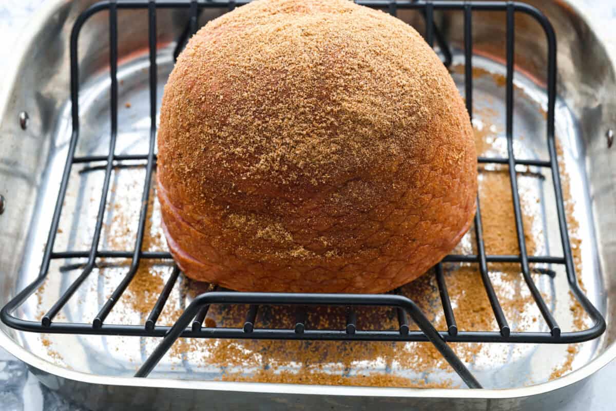 Brown Sugar Pineapple Glazed Ham is coated with a sweet and tangy spiced glaze that caramelizes on the edge of the ham. It's covered in juicy pineapple rings and maraschino cherries that add a tangy sweetness and juices to the succulent ham. Angle shot of ham covered in brown sugar and spices in a roasting pan.