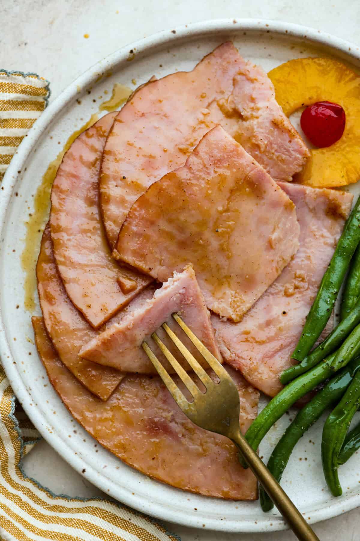 Brown Sugar Pineapple Glazed Ham is coated with a sweet and tangy spiced glaze that caramelizes on the edge of the ham. It's covered in juicy pineapple rings and maraschino cherries that add a tangy sweetness and juices to the succulent ham. Close up shot of plated brown sugar pineapple glazed ham next to green beans, a pineapple ring and a cherry.