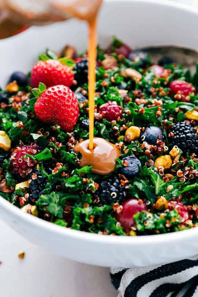 Berry, quinoa, and kale salad in a white bowl with strawberries on top and dressing being poured.