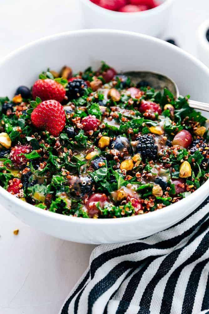 Berry, quinoa, and kale salad in a white bowl with a metal spoon, placed on a black and white stripped cloth.