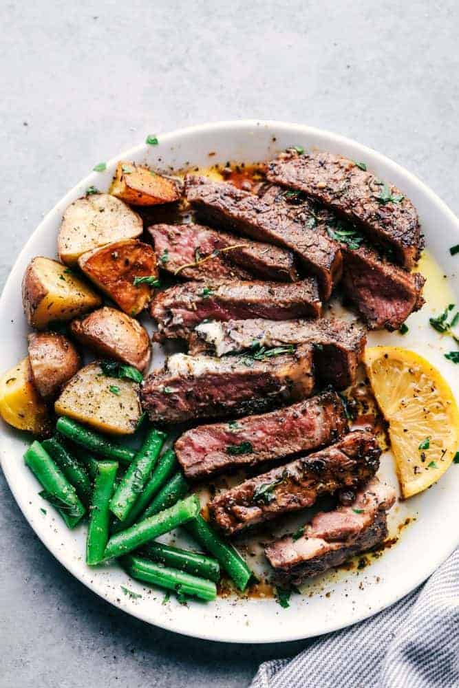 Marinated and cooked steak with cooked potatoes, green beans and sliced lemons on a white plate. 