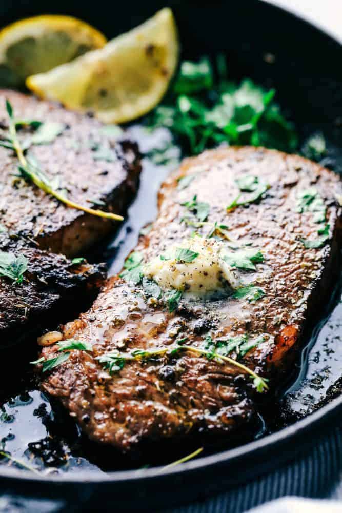 Cooked steak in a skillet with marinade, lemons and garnish on top.
