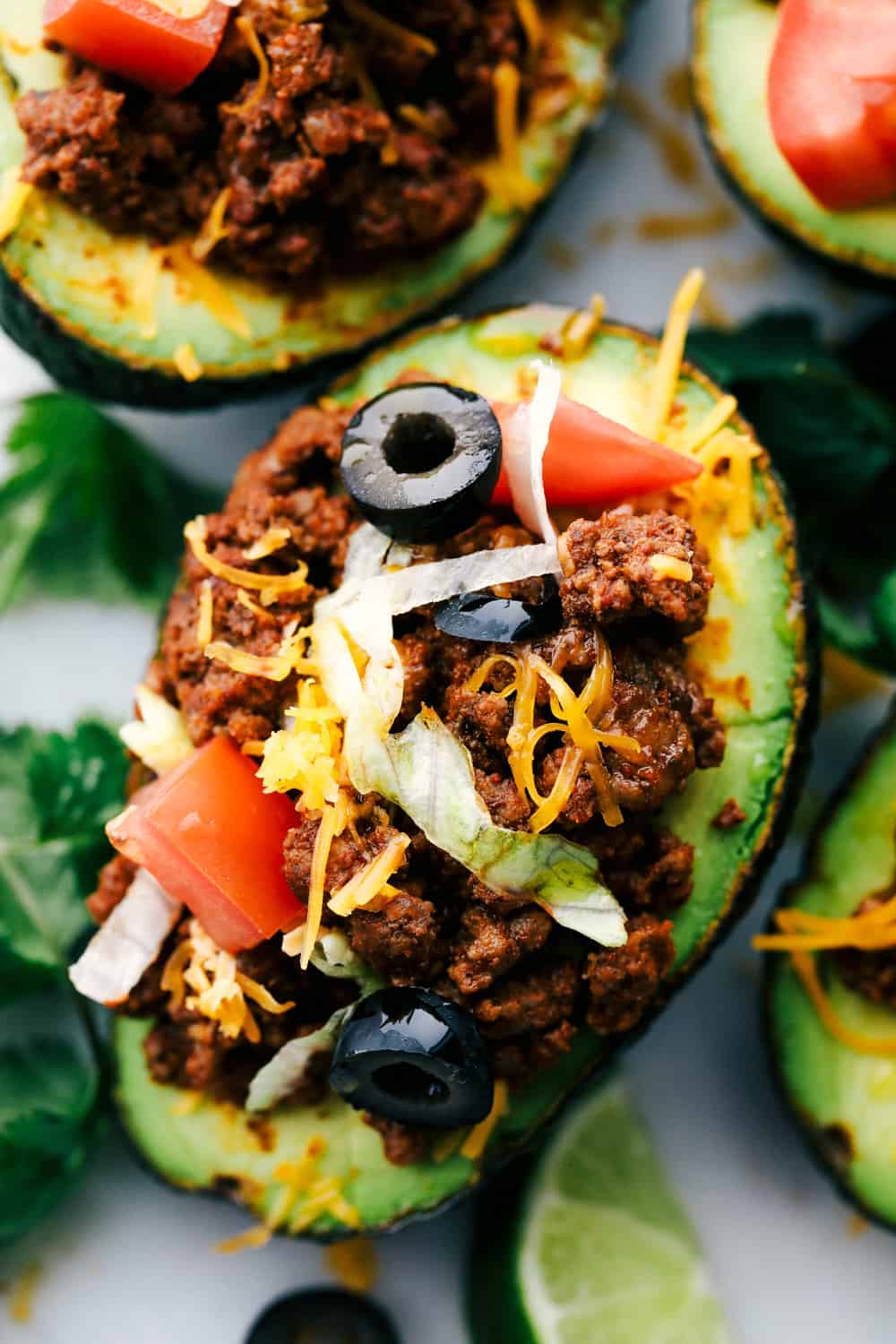 Close over head view of taco stuffed avocados on a white marble slab. The avocados are garnished with tomatoes, olives, cheese, and cilantro.