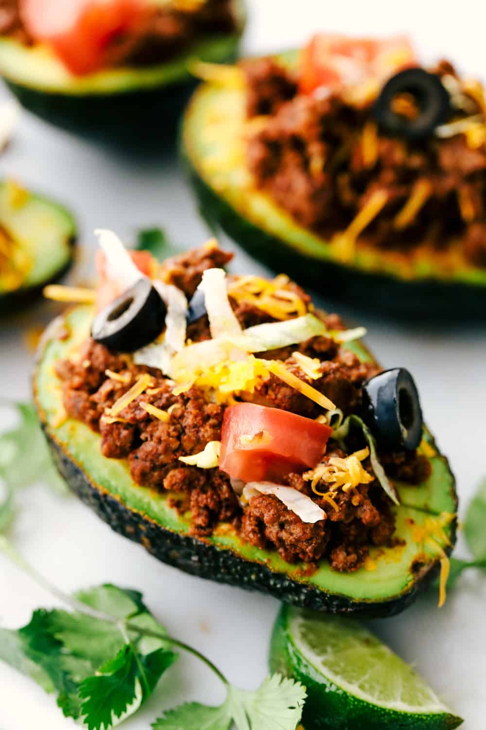 Side view of a taco stuffed avocado. The avocado is garnished with tomatoes, olives, cheese, cilantro, and a lime wedge.