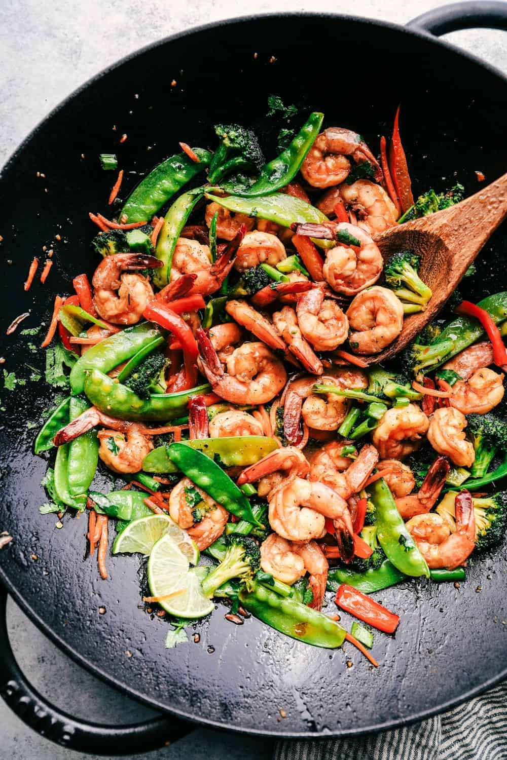 Shrimp stir fry in a large wok with lime slices and a wooden spoon.