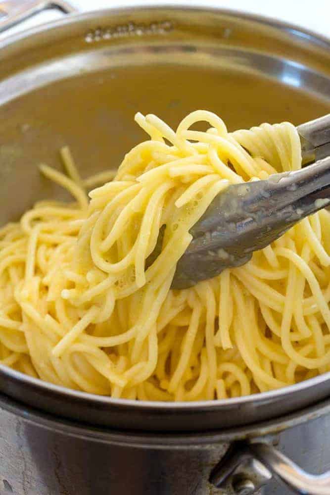Metal tongs picking spaghetti noodles out of a metal bowl.