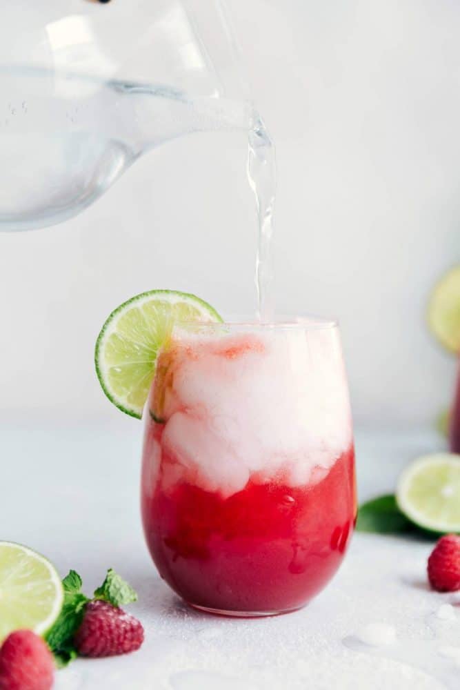 Raspberry Limeade in a clear glass with fresh water being poured into the glass. There is a sliced lime on the glass as well. 