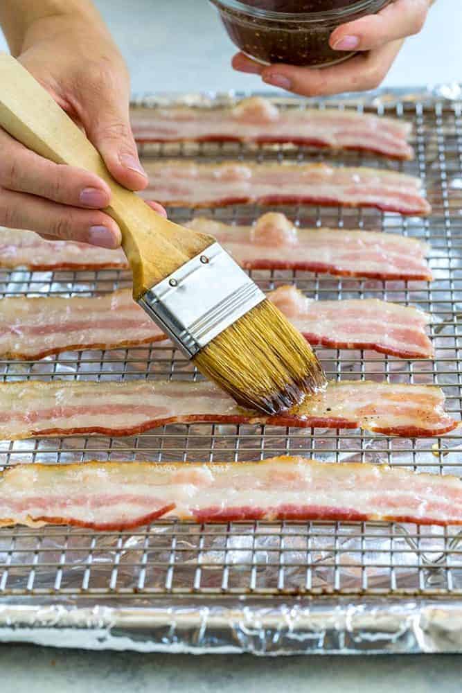 Hands brushing maple syrup glaze on slices of bacon.