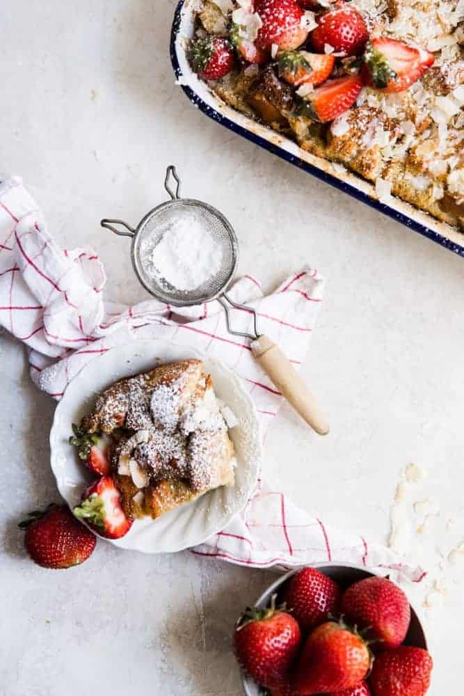 Coconut Bread Pudding on a white plate with powdered sugar and fresh strawberries in a dish. 