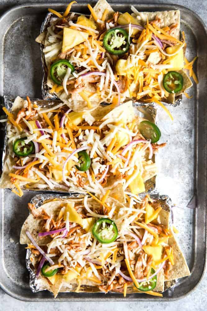 BBQ Chicken Nachos with jalapeños on top. The small aluminum foil trays are placed on a larger cooking sheet.