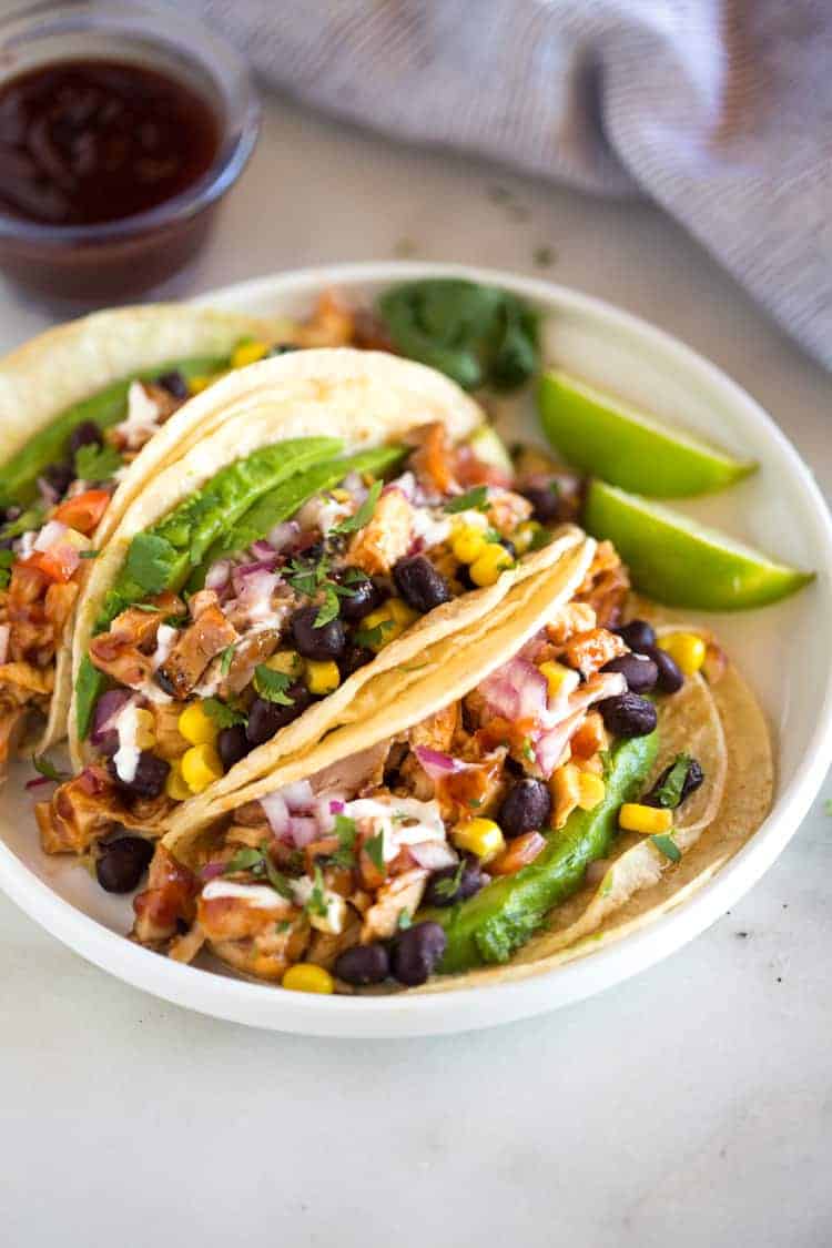 Grilled BBQ Chicken Tacos topped with corn, black beans, avocado, onion and tomatoes, served on a white plate with a small container of extra bbq sauce in the background