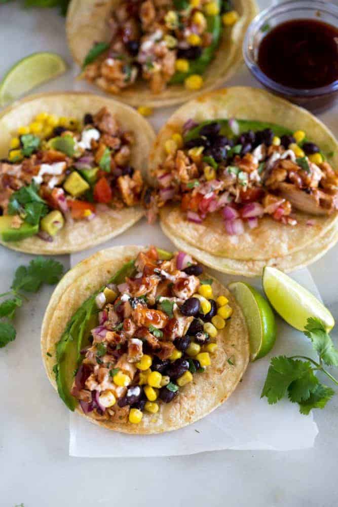 The ingredients for bbq chicken tacos piled on flat corn tortillas, including chicken, avocado, black beans, corn, and onion, with lime wedges and cilantro on the side. 