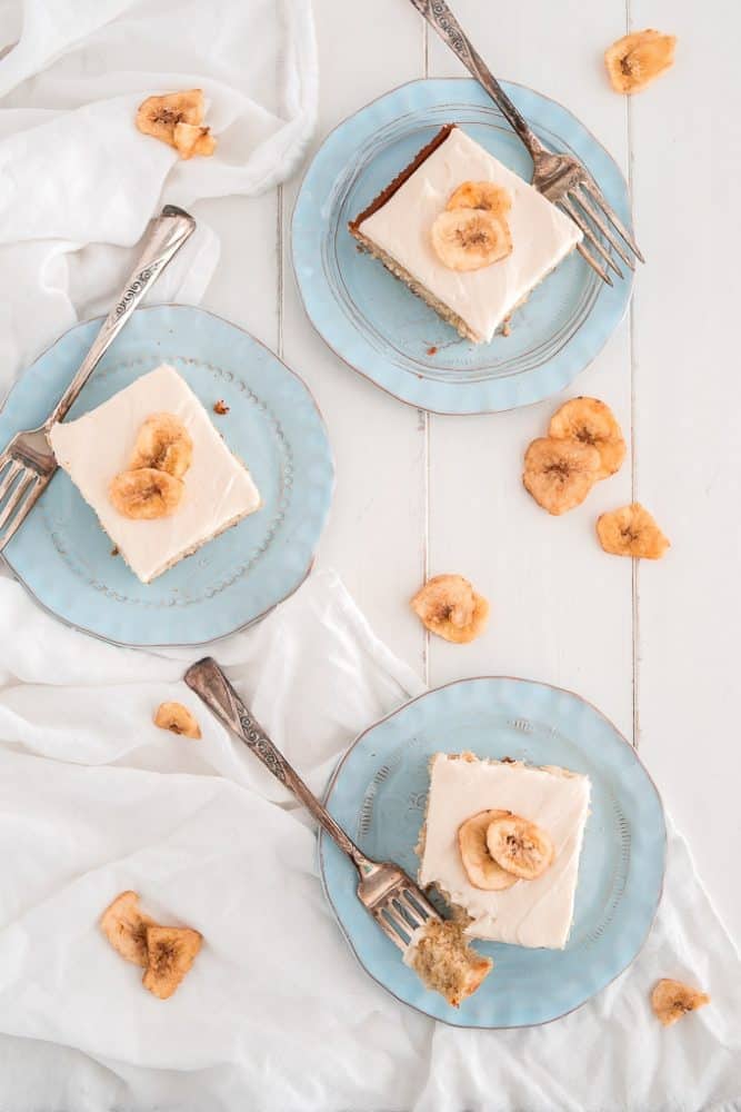 Three slices of Banana Cake with Cream Cheese Frosting on light blue plates and metal forks. There are banana chips on top of the cake slices and on the table. 