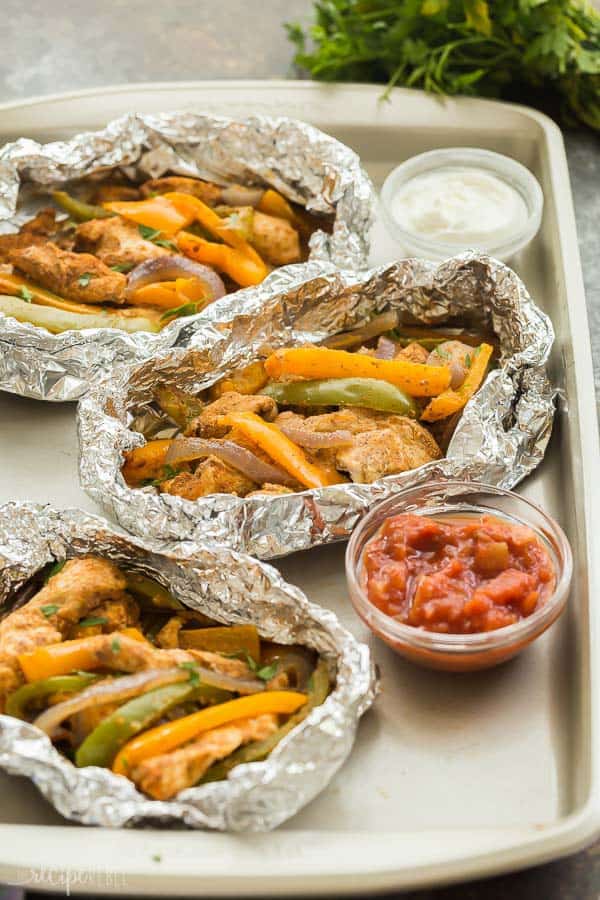 Chicken fajita foil packets on a cookie sheet with a small dish of salsa and sour cream. 
