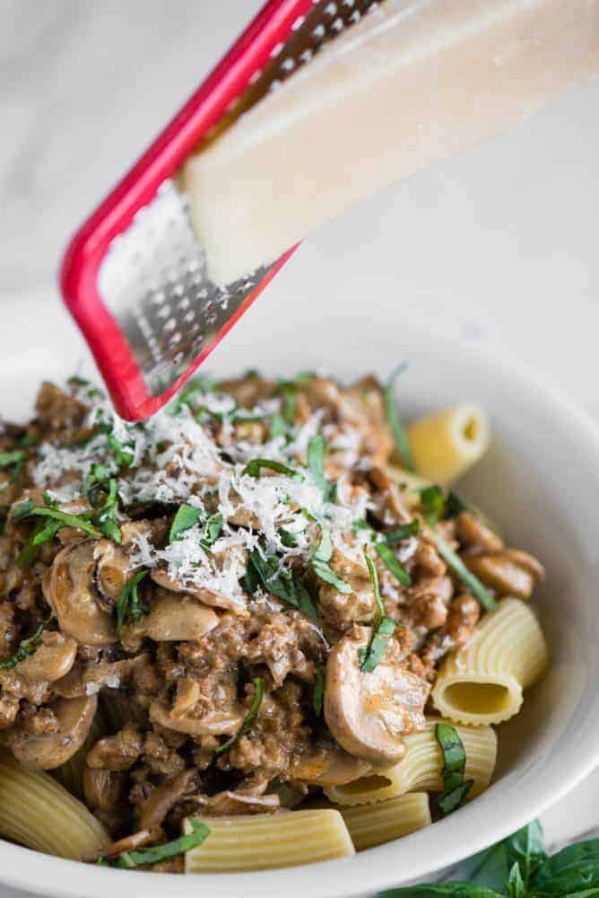 Cheese being freshly grated on top of Creamy Sausage Mushroom Rigatoni in a white bowl. 