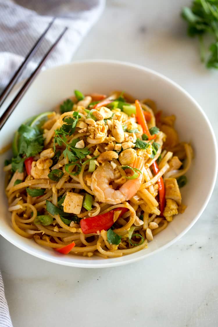 A bowl full of pad thai with rice noodles, shrimp, chicken, tofu, green onion, peanuts and scrambled egg.