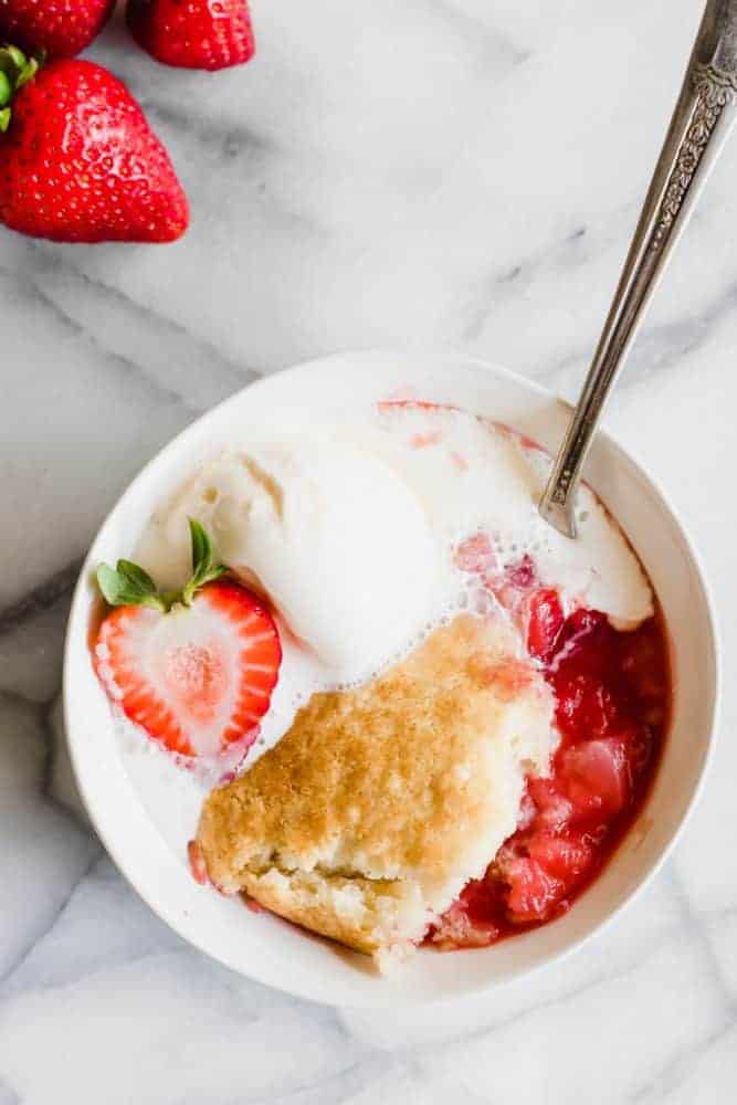 Strawberry Cobbler in a white cup with a metal spoon. There are whole fresh strawberries on the side. 