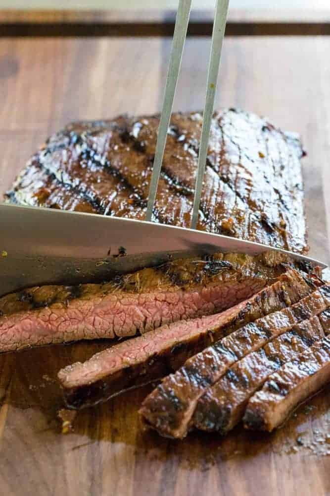 Knife slicing flank steak into strips on a cutting board.