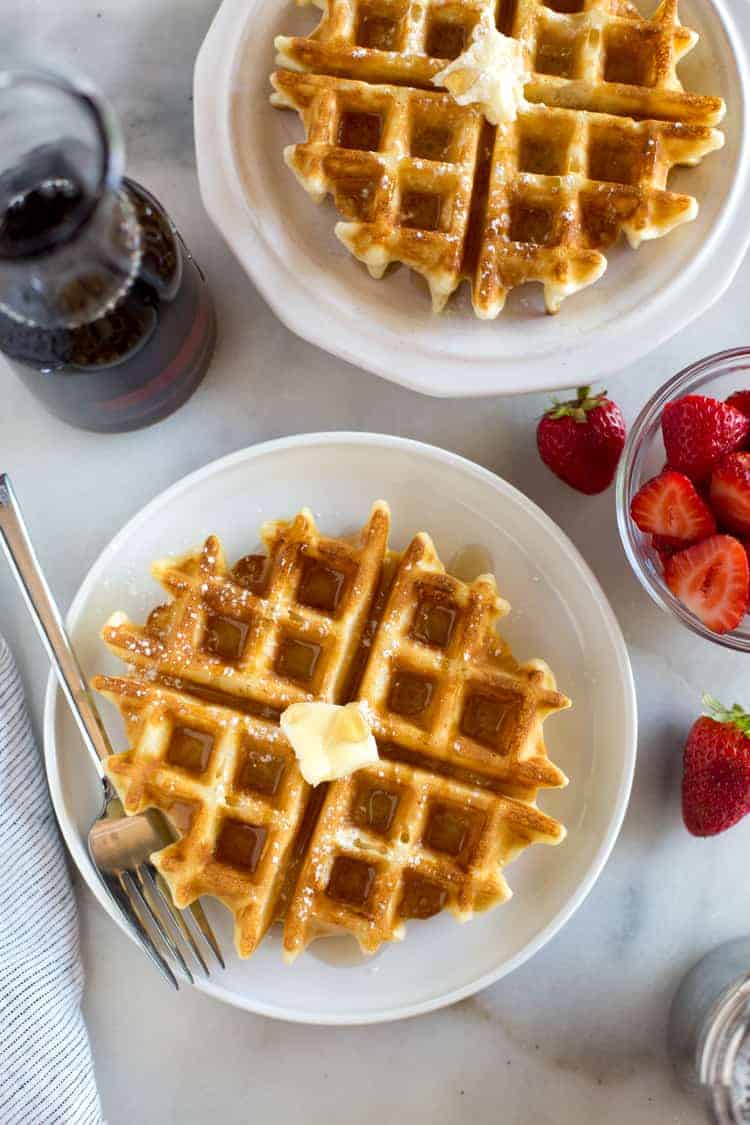 Overhead photo of two plates with belgian waffles on them, and syrup and strawberries on the side.