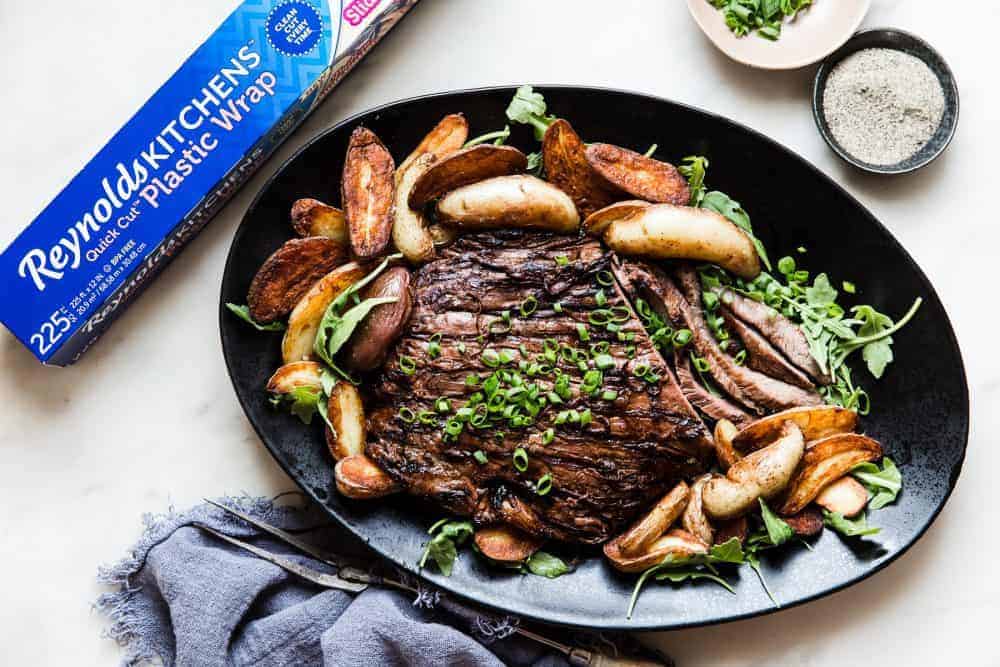 Brown Sugar Garlic Flank Steak on a black serving platter with a box of Reynolds Plastic Wrap next to it. 