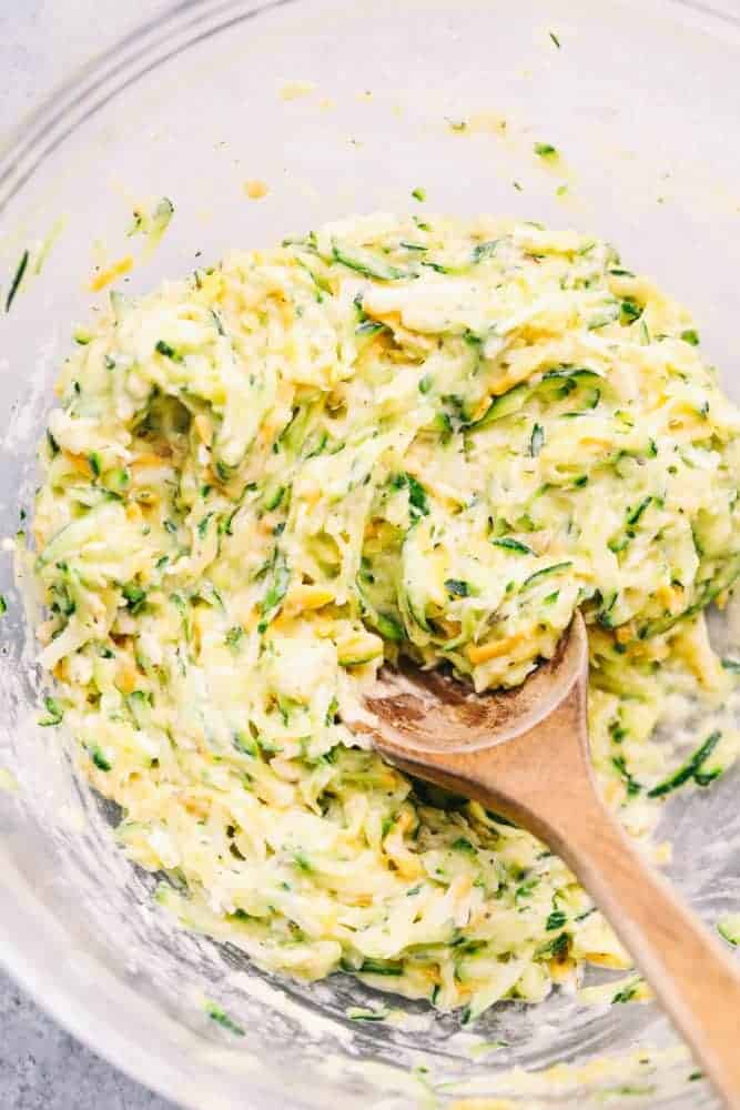 Mix for Cheesy Zucchini Fritters in a clear bowl with a wooden spoon. 