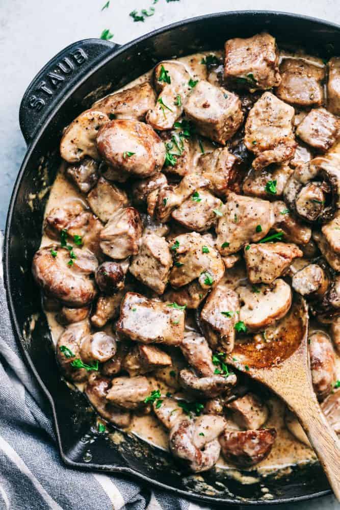 Creamy garlic streak bites with mushrooms cooking in a staub pan with a wooden spoon stirring it. 