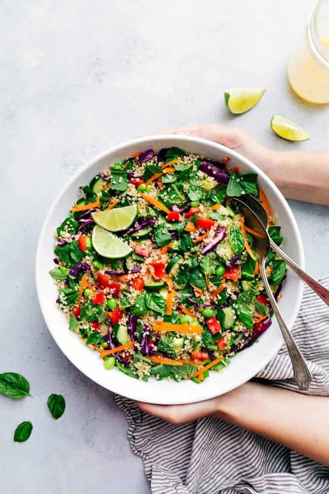 Thailändischer Quinoa-Salat in einer weißen Servierschüssel mit einer Metallgabel und einem Löffel.  Als Beilage gibt es geschnittene Limetten. 