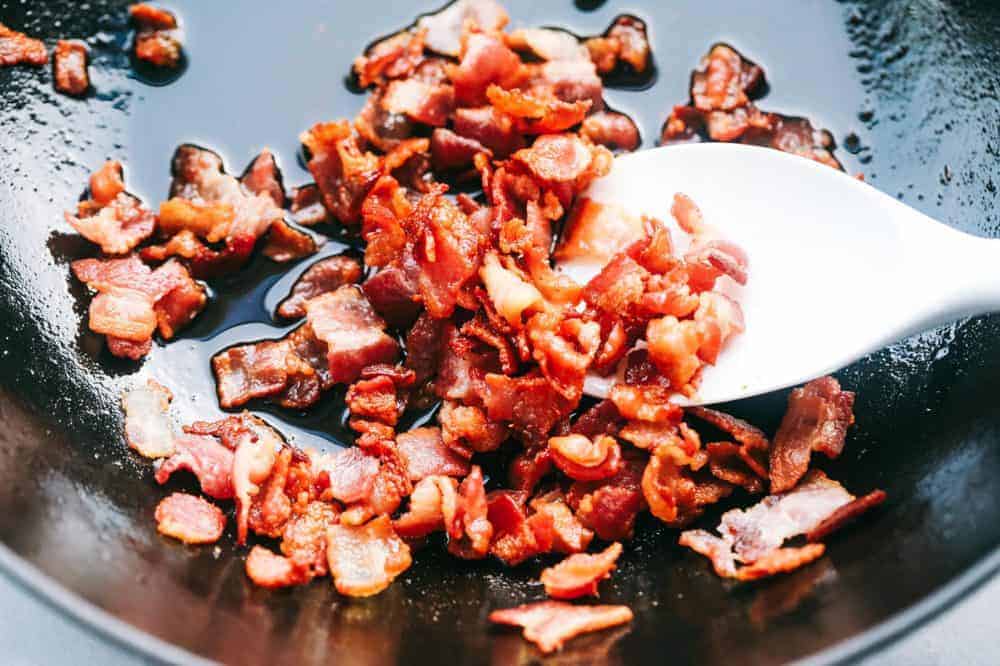 Bacon being fried in a frying pan using a wooden spoon. 