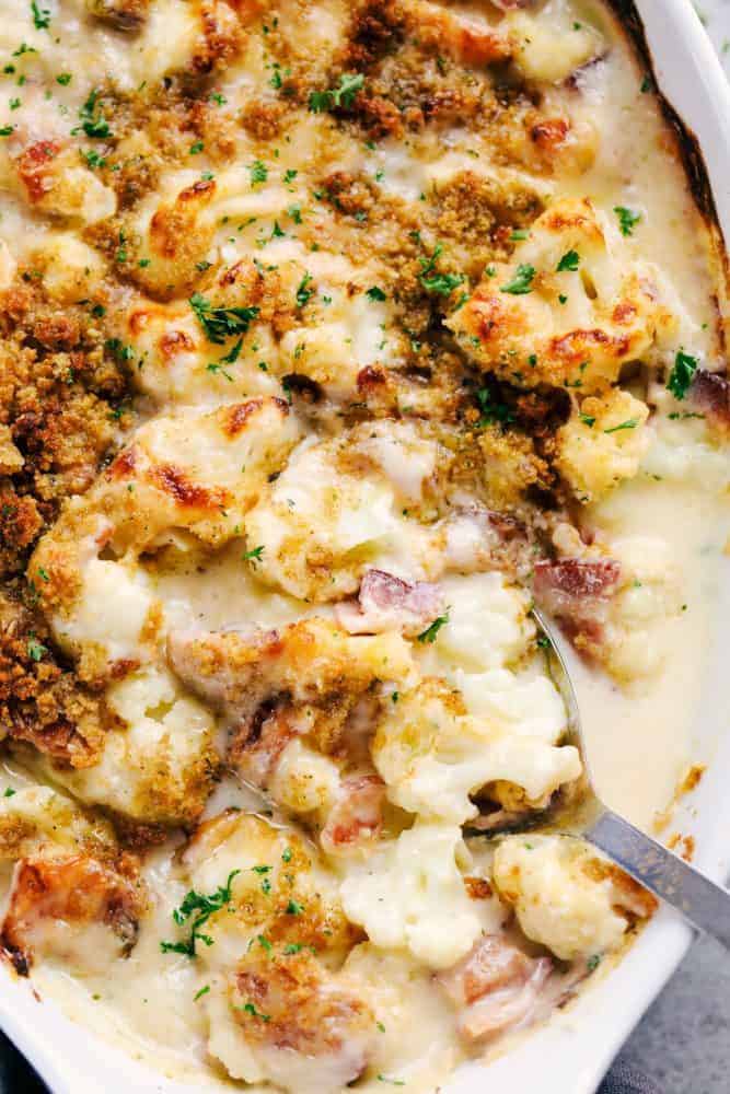 Creamy cauliflower au gratin with bacon in a white casserole dish with a silver spoon pulling up part of the cauliflower and au gratin with bacon to show the chunks.