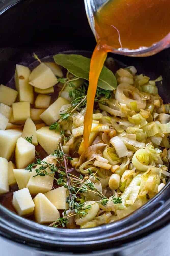 Broth being poured over leeks and potatoes inside a slow cooker.