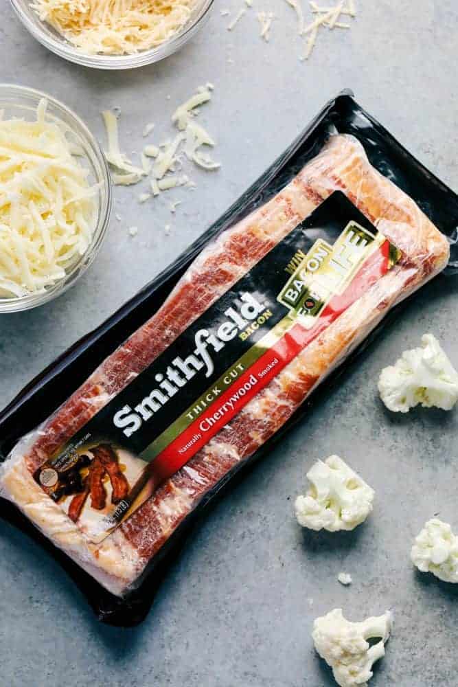 A photo of Smithfield thick cut cherrywood bacon in the middle of the photo with cut up cauliflower on the side and shredded cheese and two glass bowls.