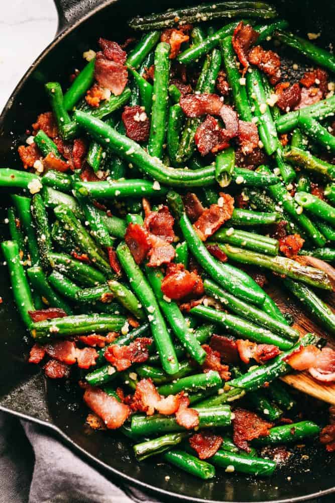 Garlic Parmesan green beans with bacon and a black skillet with the wooden spoon stirring.