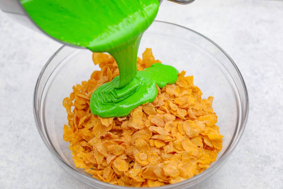 Angle shot of someone pouring the green marshmallow mixture into the cornflakes in a glass bowl. 