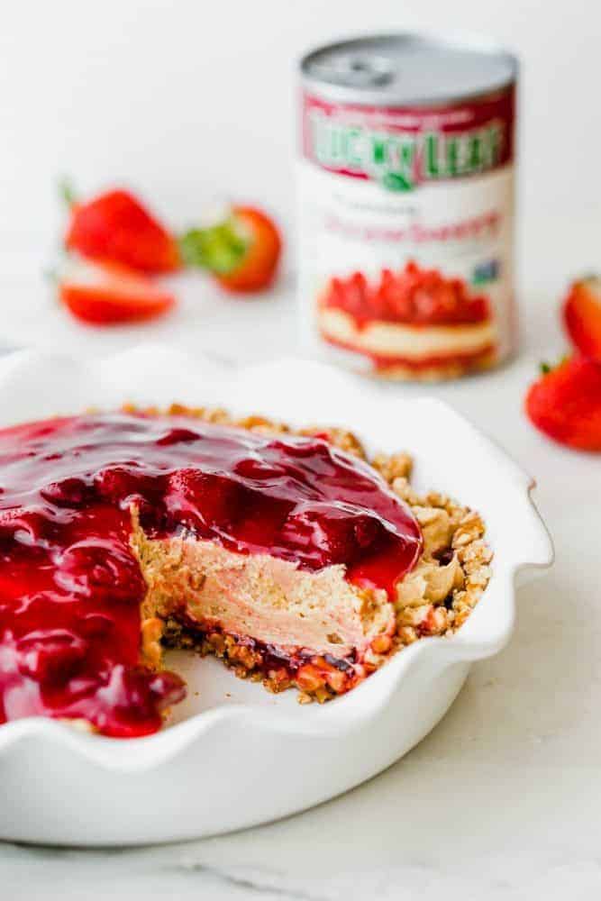 Strawberry peanut butter pie in a white pie pan with a slice removed. There are fresh strawberries and a can of pie filling in the background