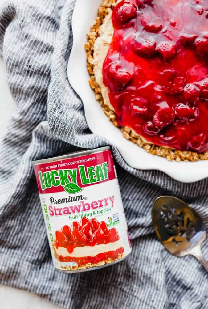 Strawberry peanut butter pie in a white pie pan with a can of Luckyleaf strawberry pie filling on the table with a metal spoon next to it. 
