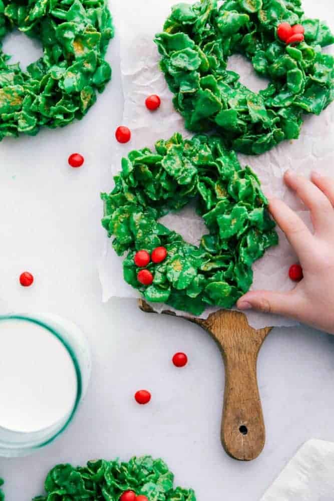 Christmas cornflake wreaths on parchment paper on a cutting board with a glass of milk on the side. There is a hand in the photo to help with size.