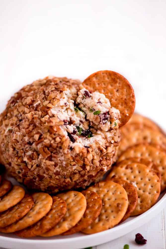 Cranberry pecan cheeseball centered in the middle of a white serving dish with crackers all around it and one crackers deck into the cranberry pecan cheese ball.