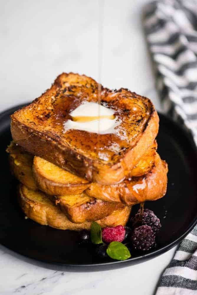 Play French toast act on top of each other on a black plate with blackberries and raspberries on the side. Syrup being poured on top.