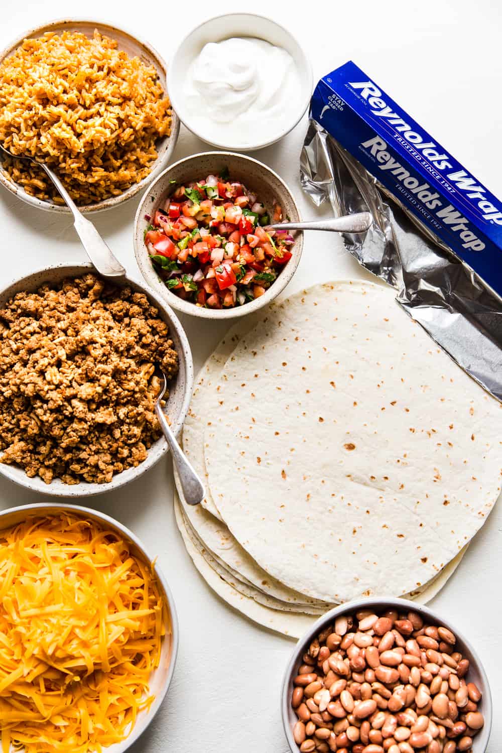 All the ingredients for a burrito. Meat, cheese, tortillas, salsa, rice, beans, aluminum foil.