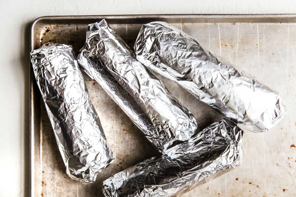 4 Burritos wrapped in aluminum foil on a baking sheet.