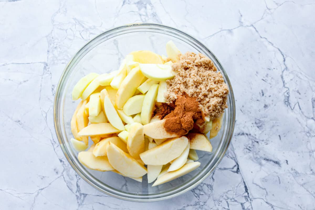 Overhead shot of peeled and sliced apples, with cinnamon, brown sugar, and lemon juice in a glass bowl. 