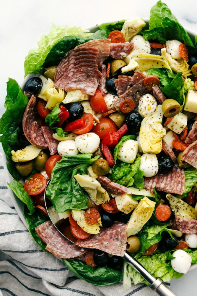 Antipasto salad with slices of meat, sliced cherry tomatoes, cheese balls, green and black olives and lettuce all in a bowl with a metal spoon.