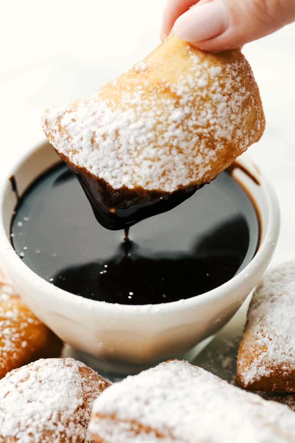A beignet covered in powder sugar then being dipped in chocolate syrup.