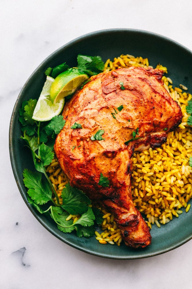 Cooked Tandoori chicken on a black plate with leaves and lime wedges on the left side and rice on the right side.