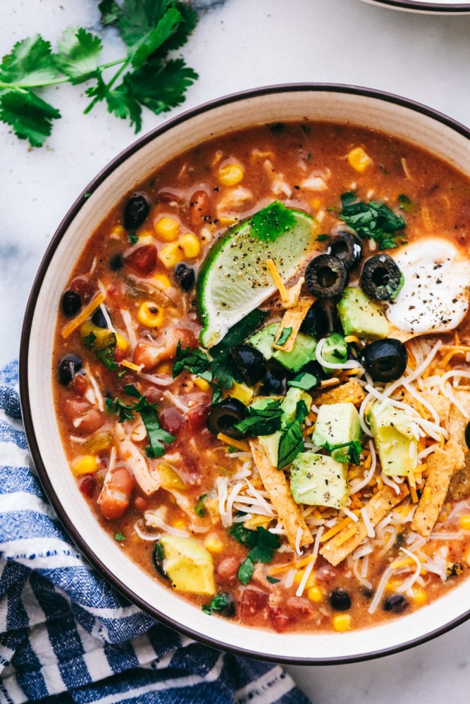 A bowl of 8 can chicken taco soup garnished with lime wedges, sour cream, sliced olives, avocado and tortilla strips. 