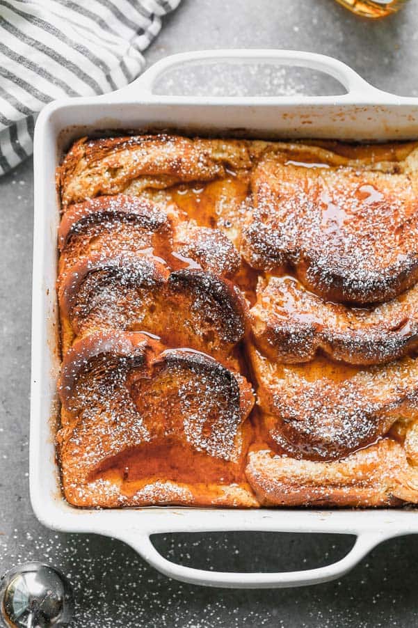 French toast in a white pan baked and syrup is drizzled over. 