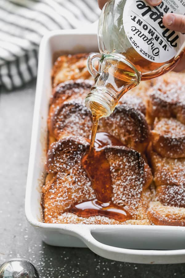 French toast baked in a white baking pan with maple syrup being poured out of a glass bottle over top of the French toast. 
